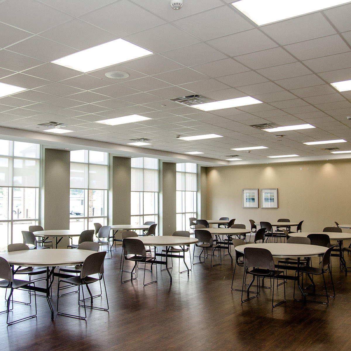 A large, open room with tall windows and a suspended grid ceiling is lit by fluorescent troffers that have been retrofitted to LED using a magnetic LED retrofit kit. Nine circular tables, each with 2 to 4 chairs are present in the room. There are two framed posters on the far wall.