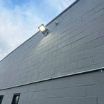 LUX LED Flood Light mounted on gray building wall shining bright under a clear sky at dusk